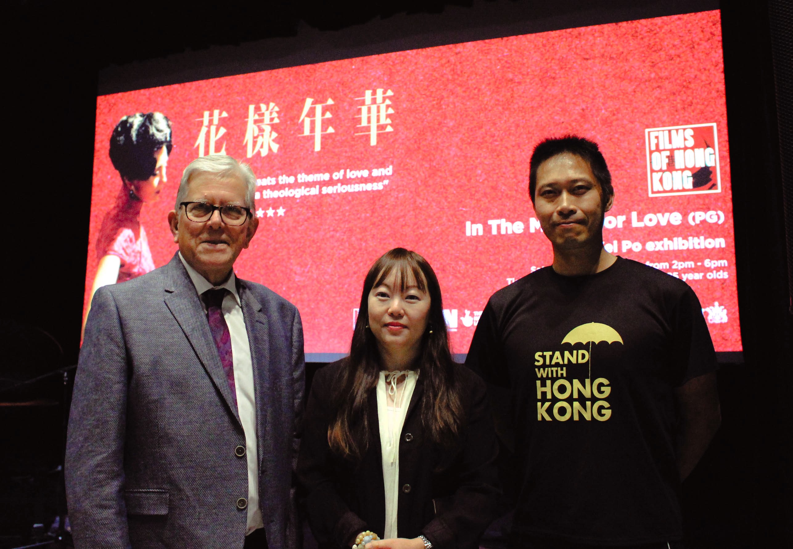 three people stood infront of a cinema screen. The Screen is promoting Hong Kong Cinema season of films.
